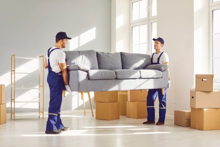 Two young delivery service workers in uniform lift heavy sofa. Men remove furniture and belongings from apartment to load in van and transport to new place. Loaders help carry couch and other things