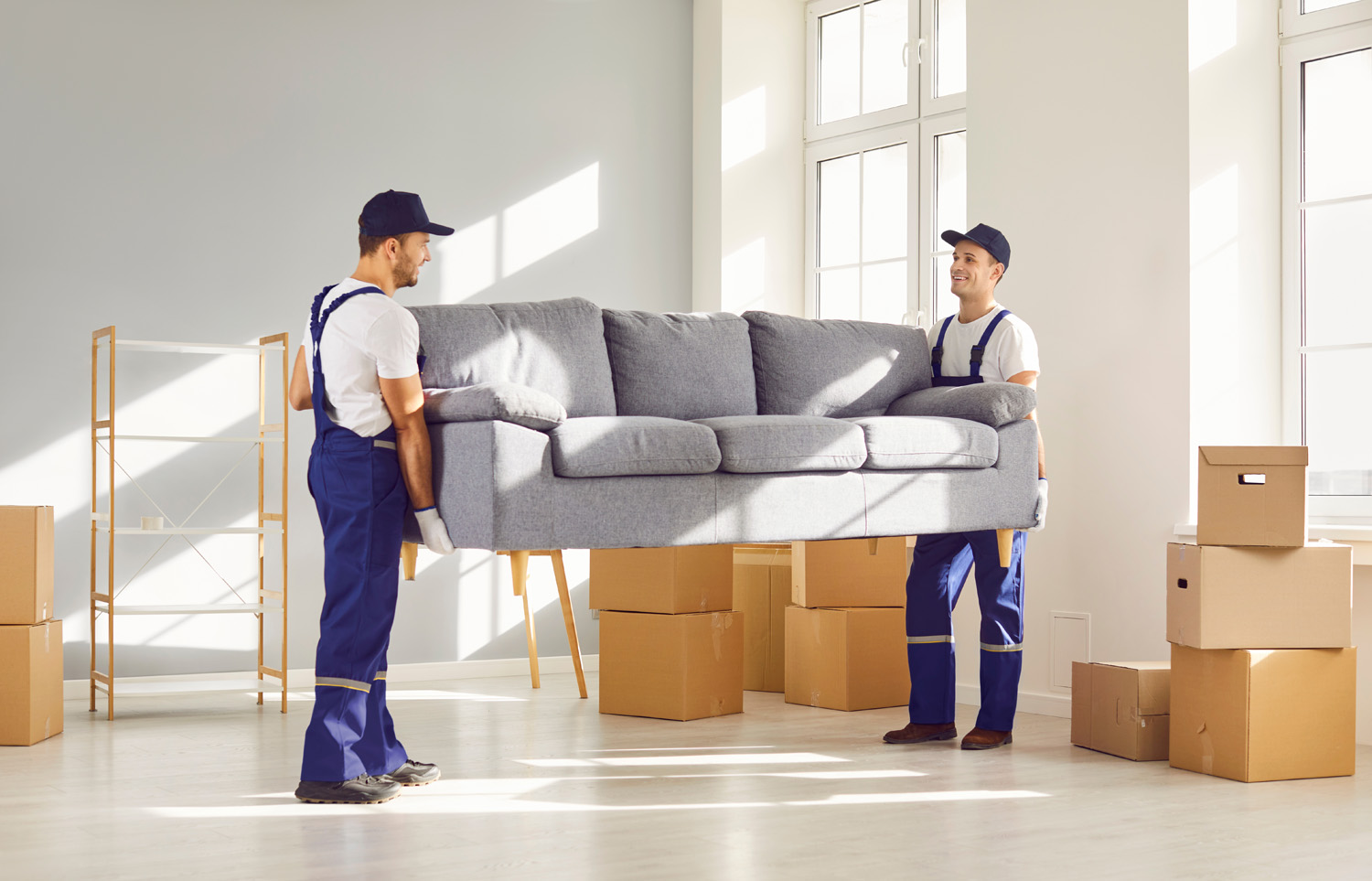Two young delivery service workers in uniform lift heavy sofa. Men remove furniture and belongings from apartment to load in van and transport to new place. Loaders help carry couch and other things