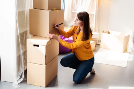 Attractive middle aged woman packer packing cardboard boxes, signing them, in apartment, moving to new house. Concept of crisis rental